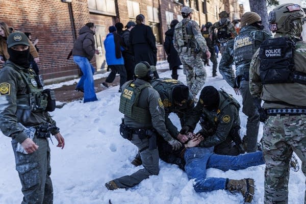 Three Border Patrol agents pin a person to the ground in the snow while surrounded by other agents.