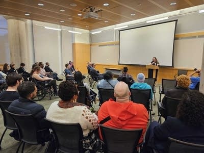 A seated crowd listens to a woman present at a podium.