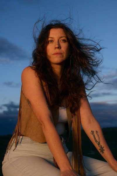 Clare Doyle wears a brown vest, white tank, light-colored denim. A dramatic blue sky is behind her.