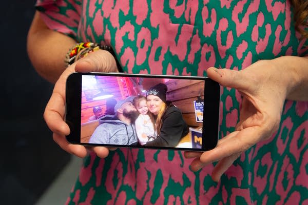 A woman displays her phone in her hands, showing a photo of a couple and their daughter