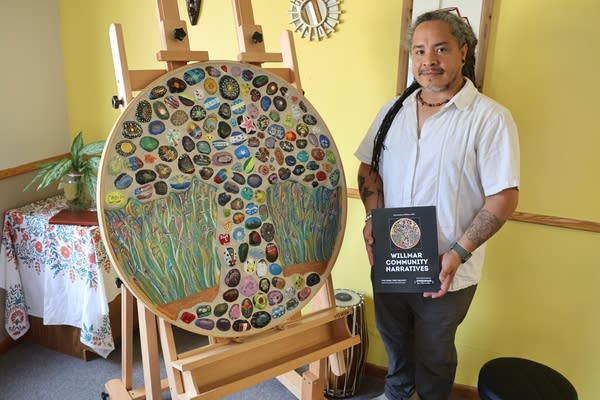 A man poses for a picture next to a round mosaic of a tree on an easel.