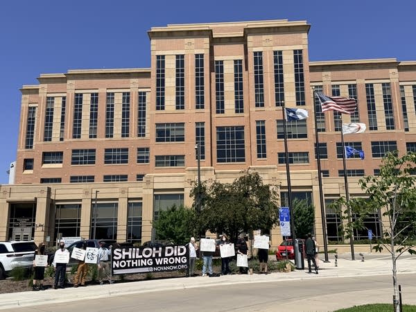 Wide shot of a courthouse. On the sidewalk in front, white supremacist protesters carry signs.