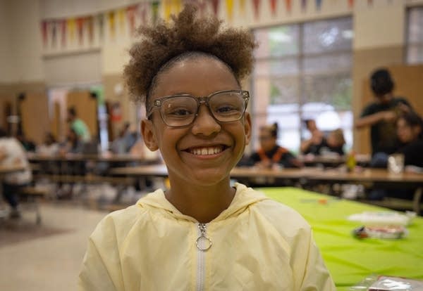 Kids smile while sitting in chairs in a cafeteria.