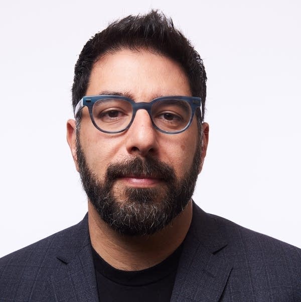Ashkan Soltani wears a navy blazer and black shirt in front of a white background