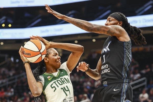A basketball player in a white uniform struggles to shoot the ball as an opposing player in a black uniform raises her arm to block.