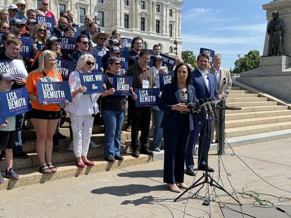 A woman speaking at Capitol with people holding signs behind her