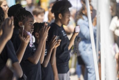 Performers in matching black shirts watch and record a dance from the sideline.