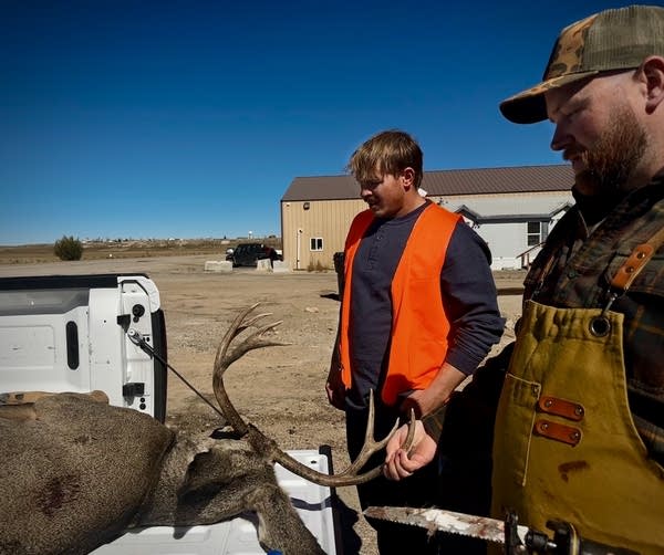 A young man in an orange vest and another man holding a saw stand in front of a dead deer in a truck bed. 