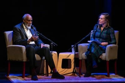 A man and woman sit in chairs on a stage, speaking into microphones.