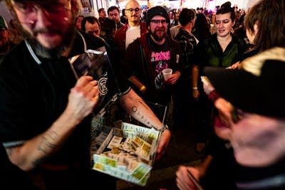 Justin Vernon wears a black shirt and cap at a bar and moves through a crowd holding pull tabs.