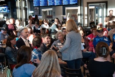 A crowd sits in a brewery, one woman stands with a microphone in hand.