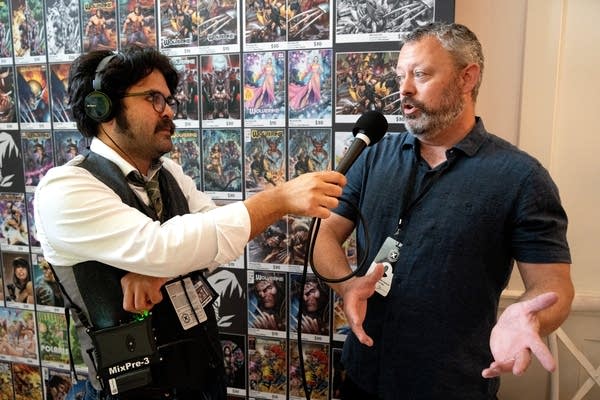 a radio reporter interviews a man in front of a wall of comics