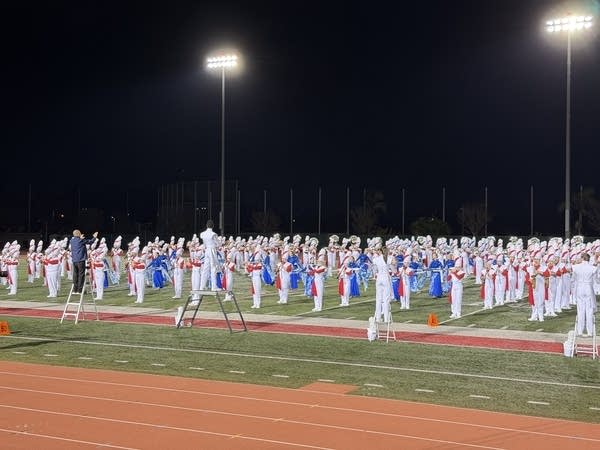 A large number of marching band players stand in formation on a football field at night.