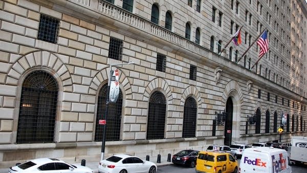 The side of the Federal Reserve Bank in New York, a stone building with American flags on flagpoles. Cars and trucks drive on the street next to the building.