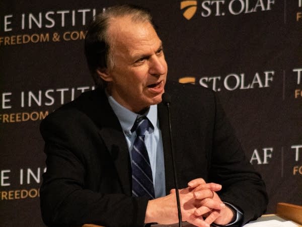 A man leans against a podium and speaks into a microphone.