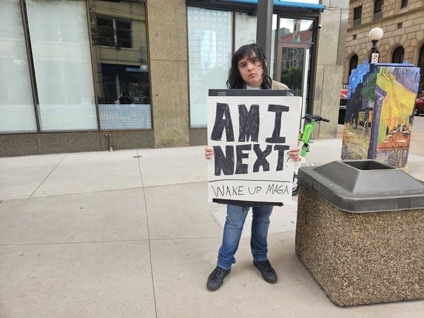 A boy poses for a photo on the sidewalk with a sign reading, "Am I next?"