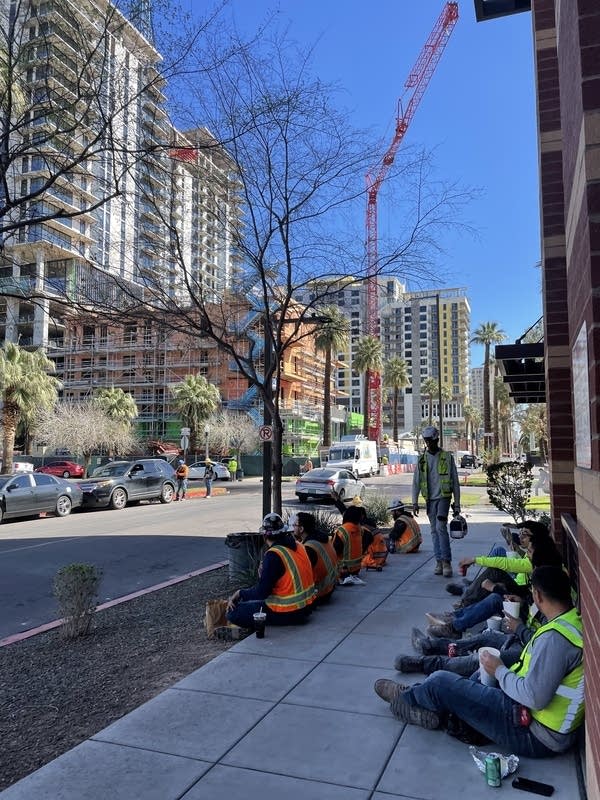Contruction workers in neon vests sit in the shade during a lunch break in downtown Phoenix, Arizona in February, 2024. In the background are new apartments under construction.