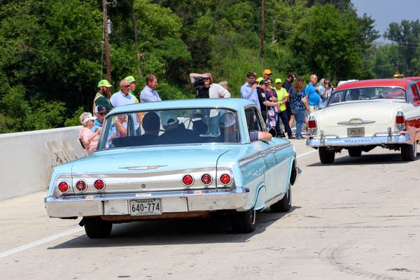 People stand on the side of a highway to watch classic cars drive past.