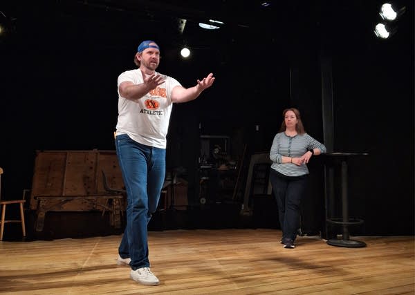 An actor on stage raises his arms during rehearsal while a woman behind him watches.