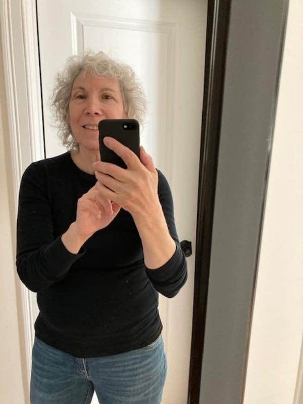 Jayne Ehrlich, a woman with gray hair in a black shirt and blue jeans, smiles for mirror selfie.