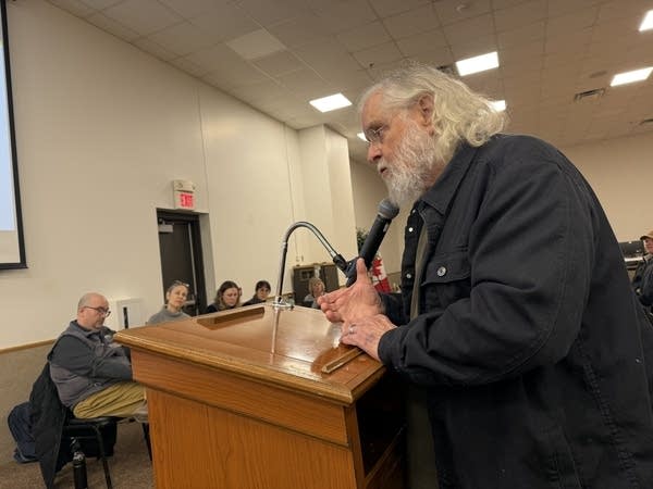 A man stands at a podium and speaks to a table of officials.