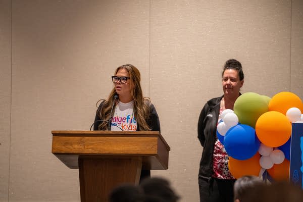 A woman speaks at a podium while another woman stands next to her, behind orange and blue balloons.