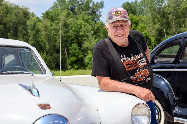A man poses for a photo by leaning on a white classic sportscar.