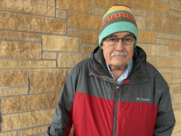 A man in a hat and winter coat poses next to a brick wall.