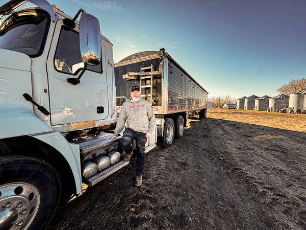 Wide-angle shot of a man in a gray hoodie standing next to a semi truck on a farm.