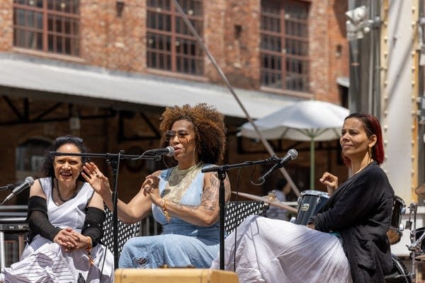 One woman speaks onstage while two other women participate