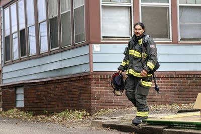 A firefighter stands next to a house, holding his helmet.