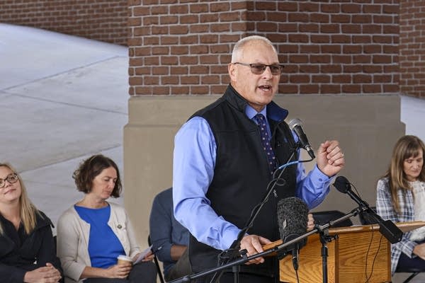 A man in a blue shirt and black vest speaks at a podium for a press conference.