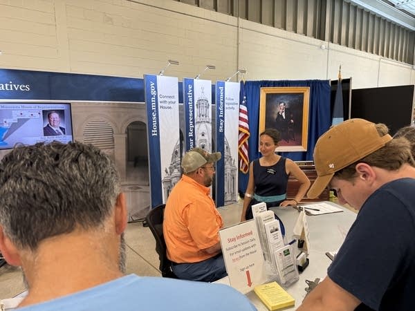 A man in a chair talks with a standing woman in front of a political display at the State Fair.