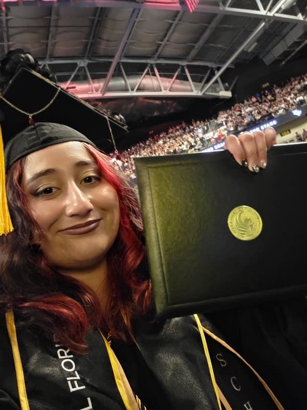 A woman poses for a selfie in a cap and gown with her diploma.