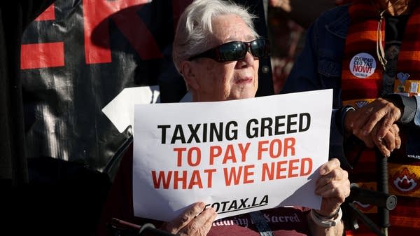 A person holds a sign that reads "Taxing greed to pay for what we need"
