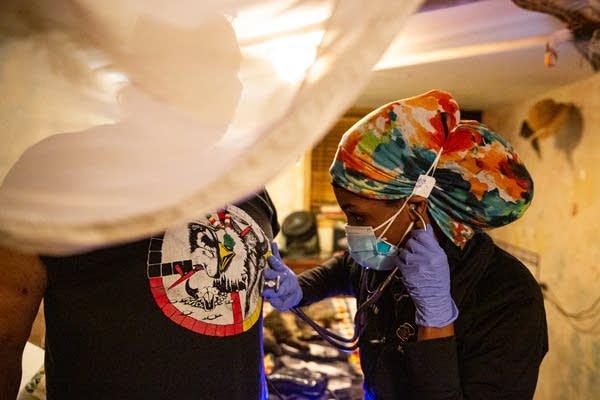 A woman in a colorful head wrap uses a sthethoscope on a man.