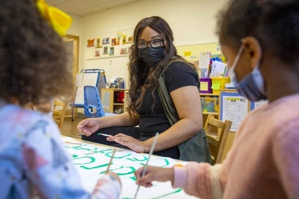 A woman with long, dark hair, a black short sleeve shirt and army green vest sits at a children's table in an early learning center. She is looking forward at two children who are blurred by the camera. They are painting letters in bright green.