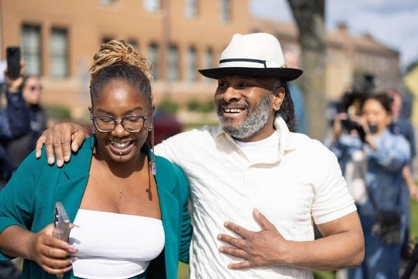 A man wearing a white shirt and hat smiles next to his daughter