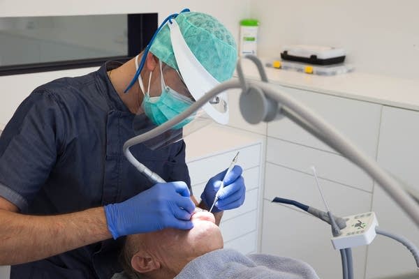 A dentist wearing a double mouth mask and a face screen treats a patient.