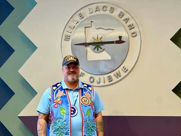 A man in a blue vest poses in front of a silver sign reading "Mille Lacs Band of Ojibwe."