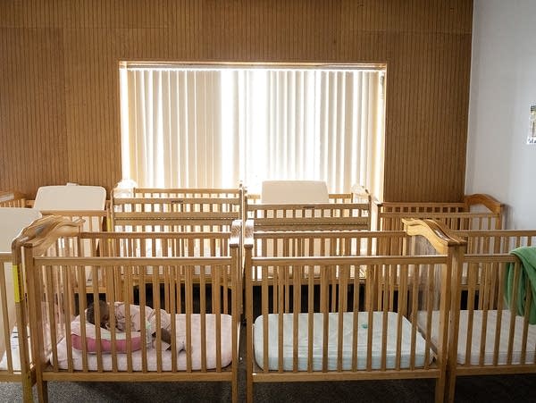 Two rows of cribs fill a room, one is occupied.