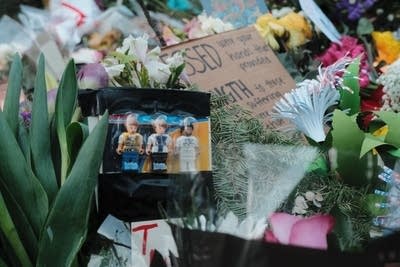 Three small toy figurines sit in a box among flowers, signs and candles.