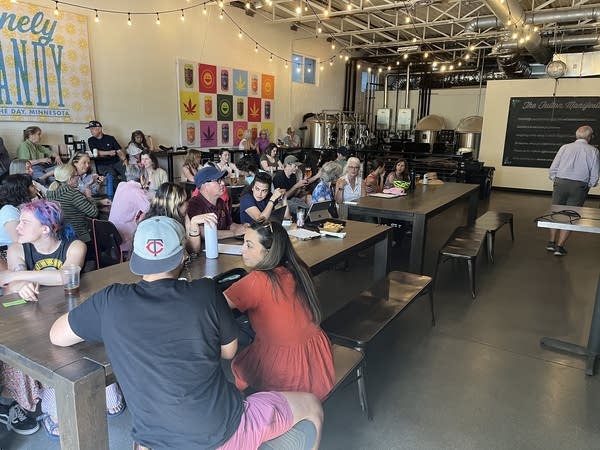 A crowd of people sit at tables to listen to a community presentation.