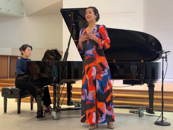 A woman in a colorful dress stands in front of a grand piano and sings.