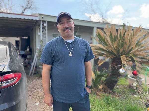 A man in a t shirt and baseball cap stands outside a home.