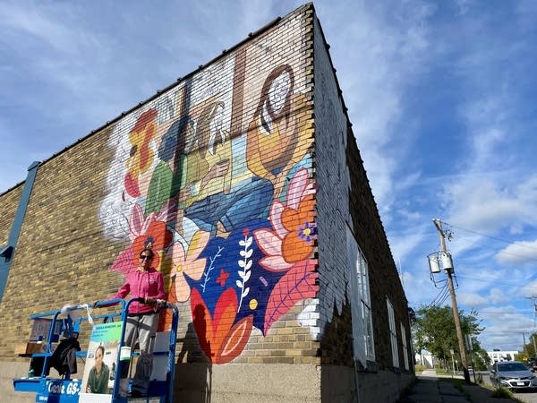 A mural artist stands on a scissor lift next to her work on the corner of a brick building.