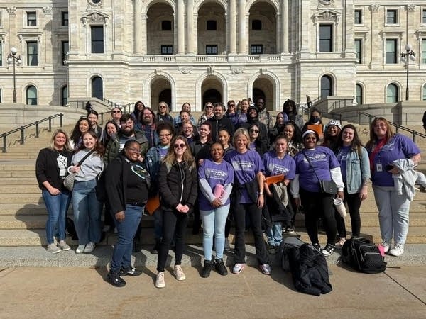 A group of people, some wearing matching purple shirts, pose outside the Minnesota State Capitol.
