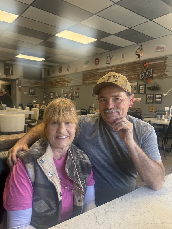 A man has his arm around the shoulder of a woman as they sit at a diner booth.