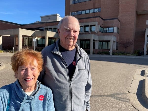 Two people smile in front of a building