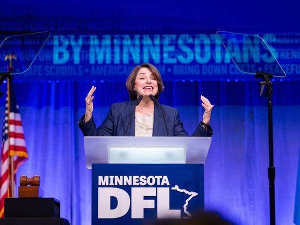 A woman raises her hands while speaking at a podium on stage.